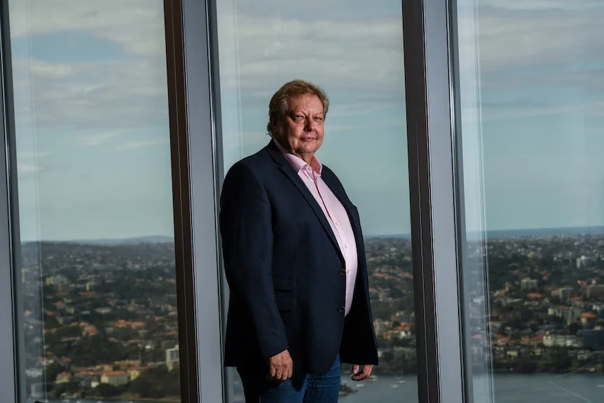 Richard White stands in front of office windows, with rooftops and trees visible behind him.