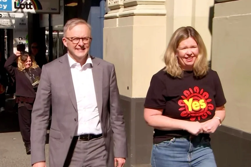 A man with glasses wearing a suit walks next to a woman wearing a t-shirt reading: Yes