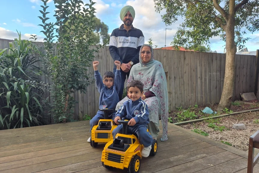 Satnam, Sikh man, wears turban, smiles, stands behind mother, head covered with stole, two children on toy scooter, smile.