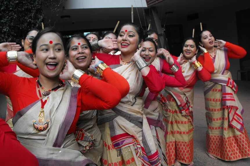 Smiling women in red blouses, cream sarees, large bindis, hair tied, have hand behind hair as they sing.