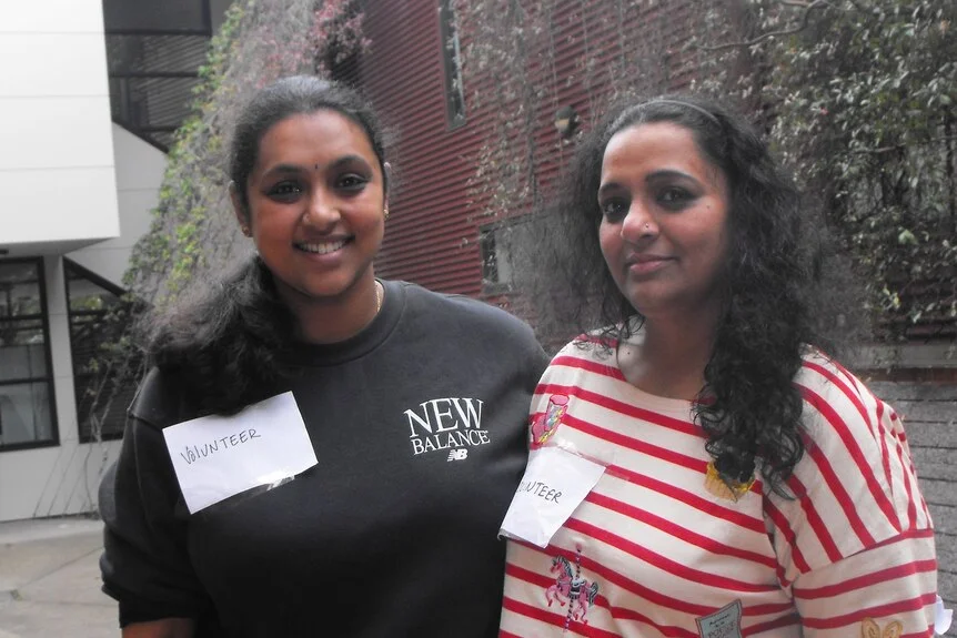 Gayathri and Varshini stand close together in a courtyard, both smile ,white paper with volunteer pinned to chest.