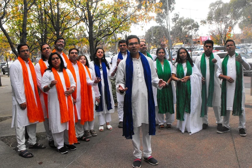 Viraj stands in front of men, women, sings, all wear kurta, white scarfs in the Indian tricolour saffron, green, white and blue.