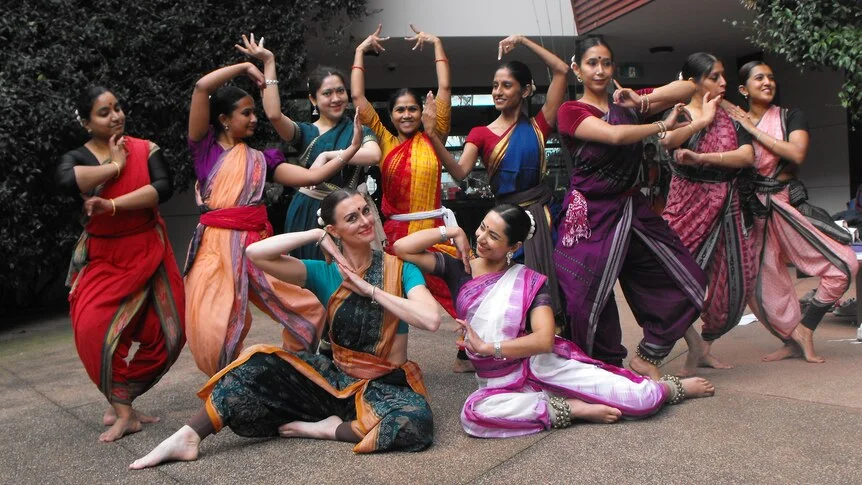 A group of women dancers dressed in a form of a saree draw poses, some stand, some sit, all smiling, in a leafy courtyard.