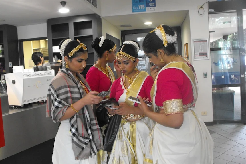 Indian classical dancers in white and gold sarees, flowers in hair, young girls, on phone.