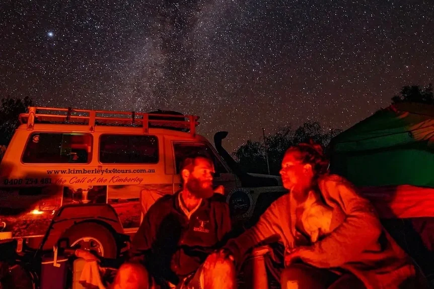 a couple sitting near their fwd, around the fire under a starry sky