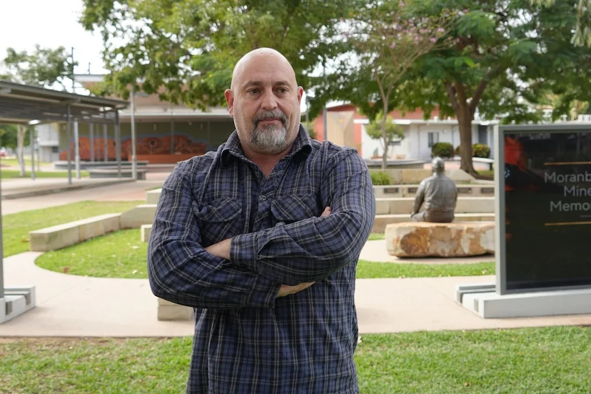 Man in flanette shirt looking off camera, arms crossed, sandstone wall with plaques of names behind