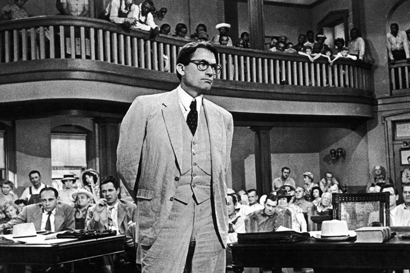 A black and white still featuring a man in a three-piece suit and thick-rimmed black glasses looking pensive in a courtroom.