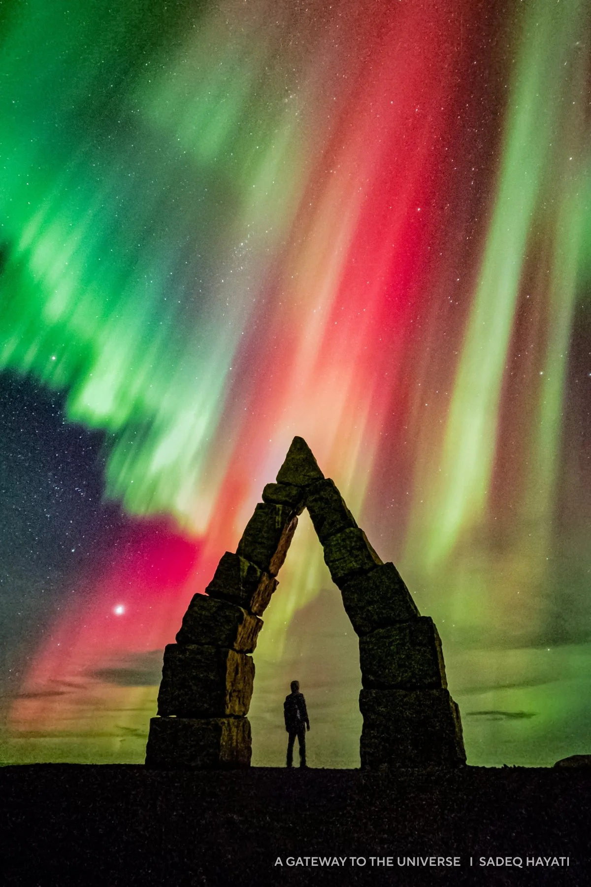 A person stands beneath a stone arch with vivid green and red auroras illuminating the 2025 night sky, creating one of the year's most captivating images.