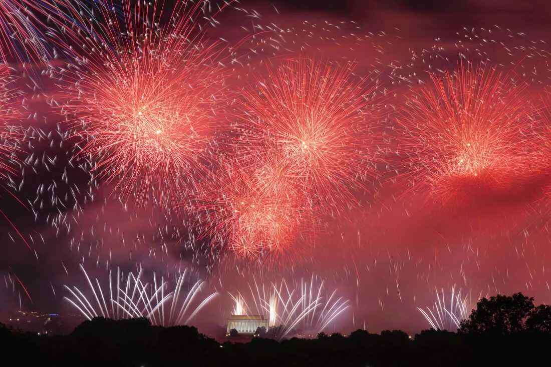 Red fireworks burst in the night sky above a lit building, with white streaks rising from the ground like a power aurora, silhouetted trees framing the vibrant display.
