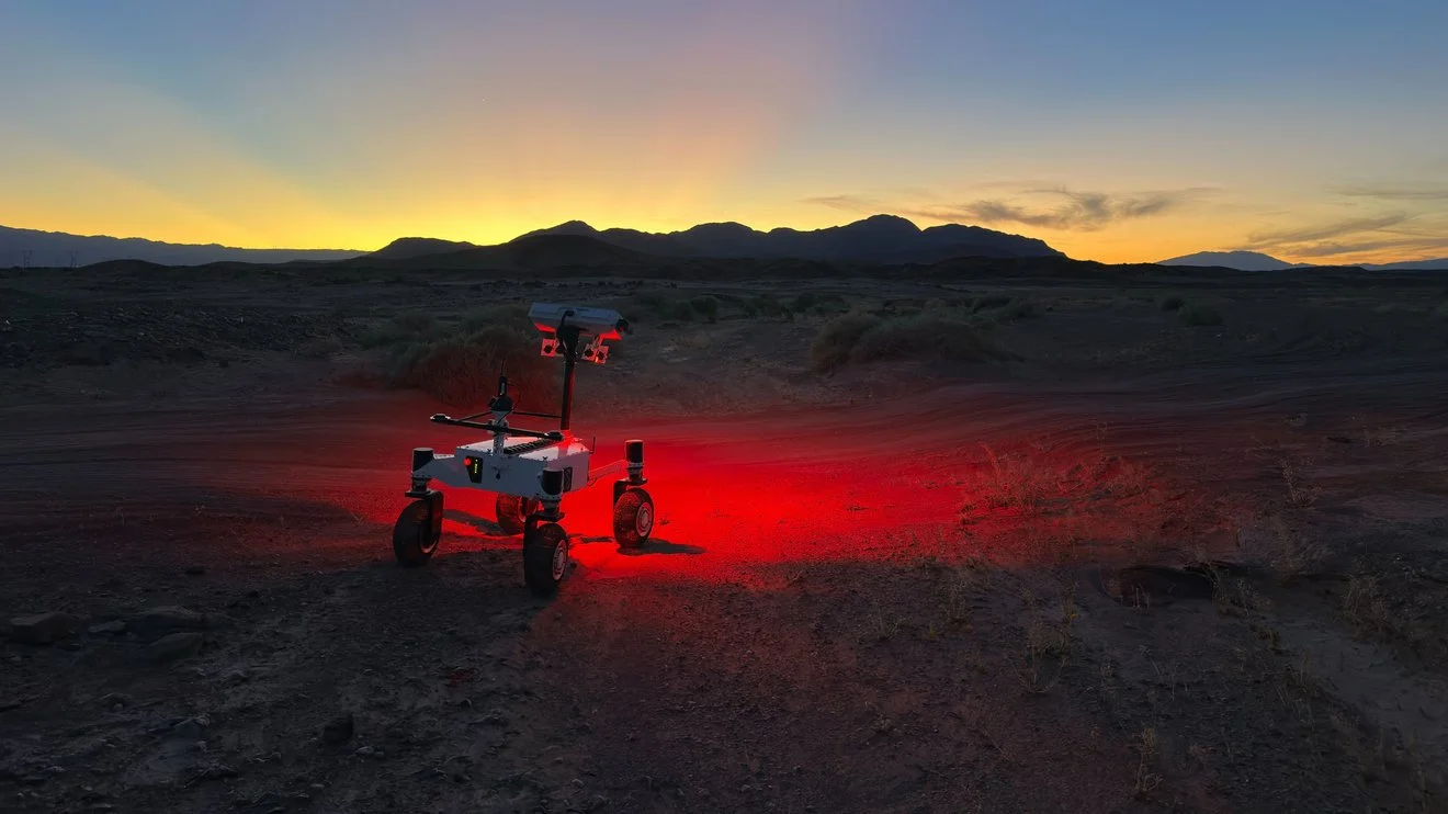 a small, four-wheeled rover shines a red light on a dark desert ground and the sun sets behind silhouetted hills in the background.