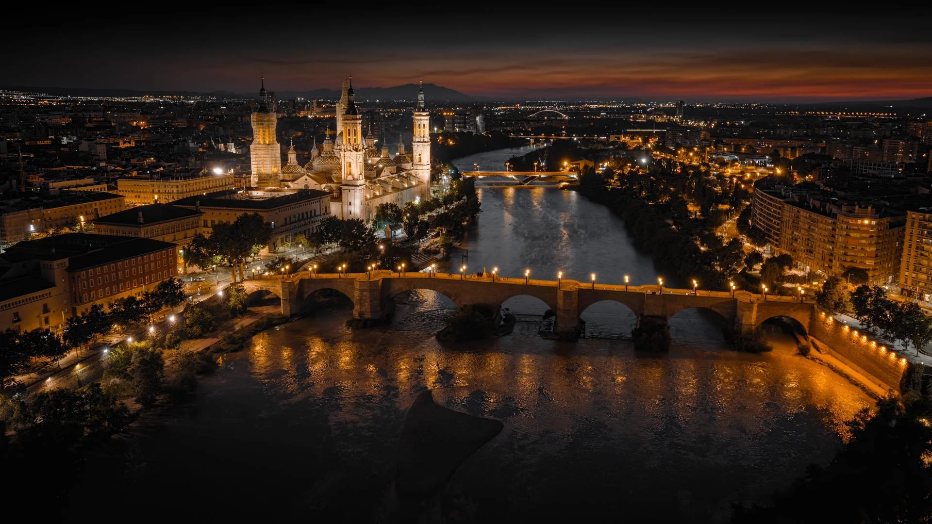A cityscape at dusk featuring a brightly lit cathedral, stone bridge, and river reflecting golden lights, with buildings and distant mountains under a dramatic, darkening sky.