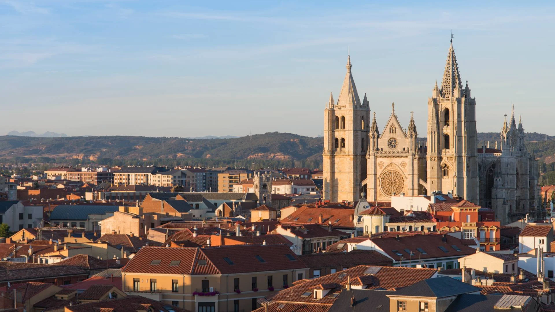 A panoramic view of a city with red-roofed buildings and the León Cathedral, a Gothic church with tall spires and ornate details, set against distant hills under a clear blue sky.