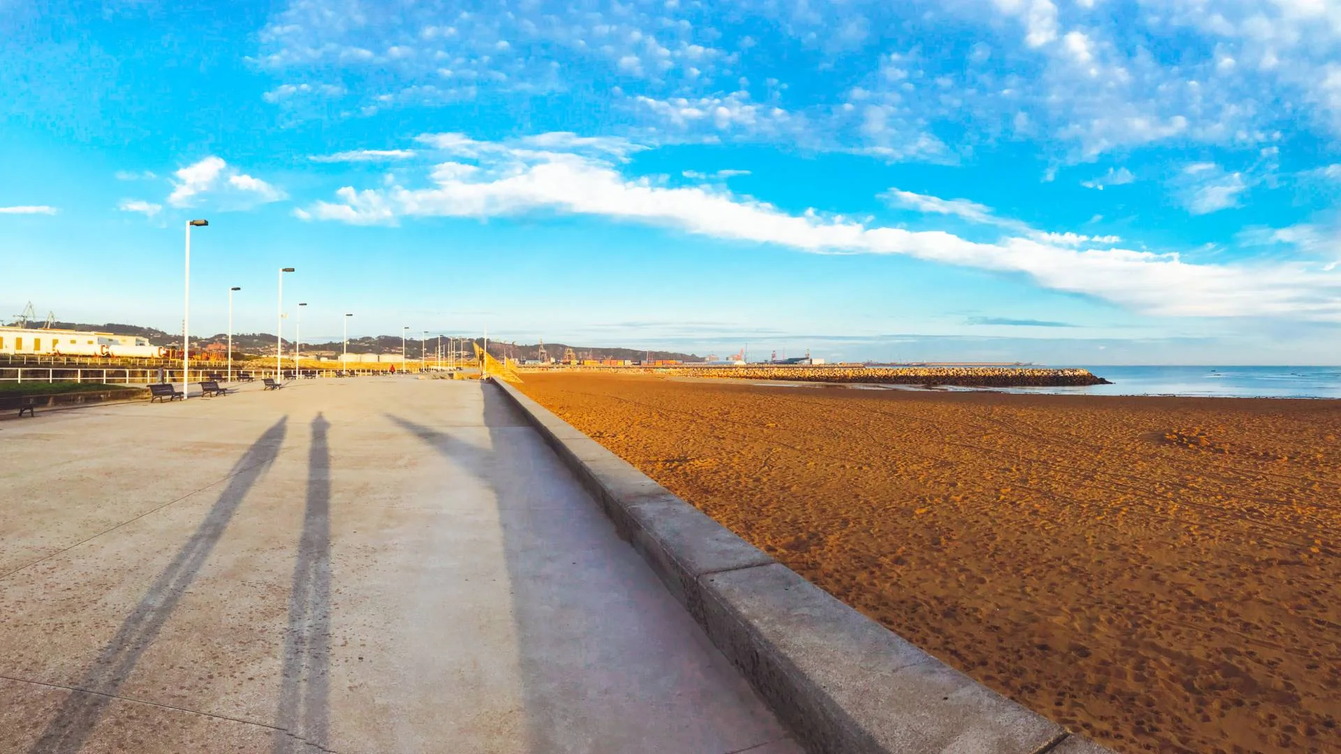 On the right is a wide sandy beach under a clear blue sky dotted with scattered clouds. On the left is a wide concrete walkway lined with lampposts and some buildings in the distance.