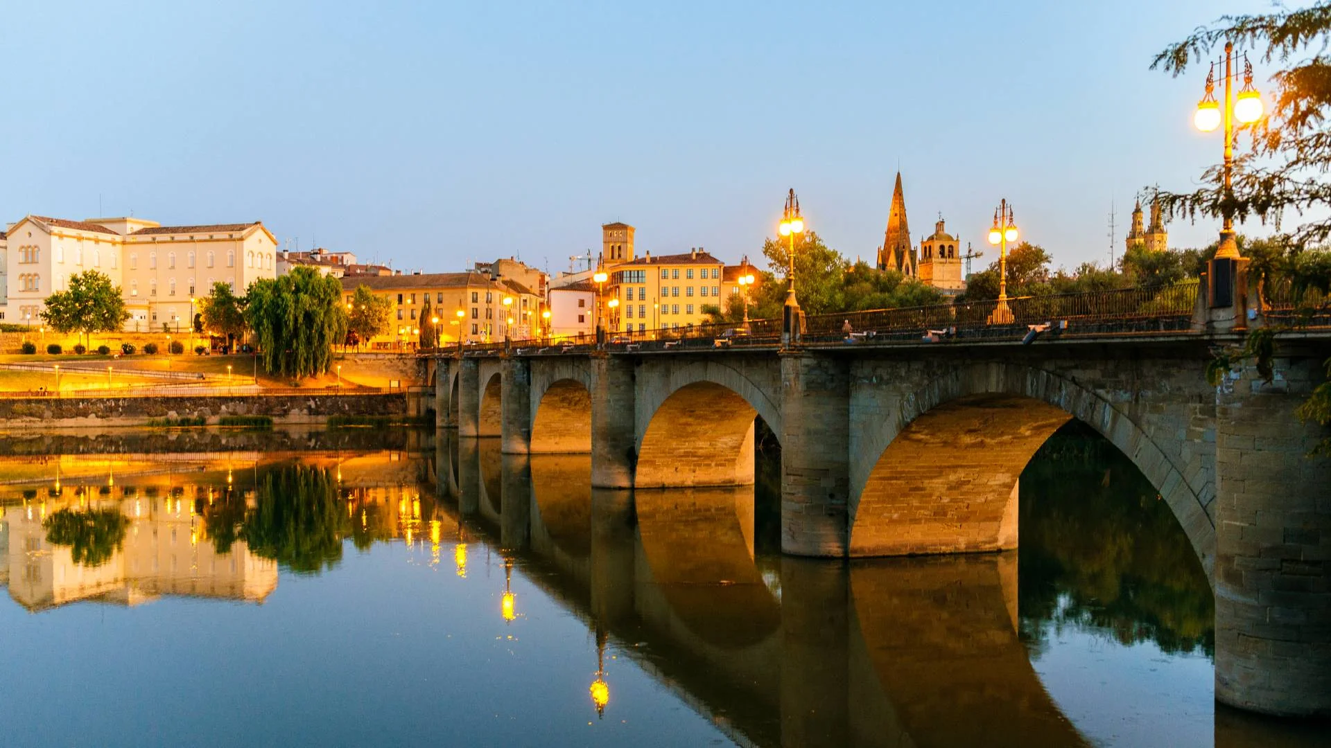 A stone bridge with arches spans a calm river at dusk, reflecting city buildings, trees, and church spires illuminated by warm streetlights against a clear evening sky.