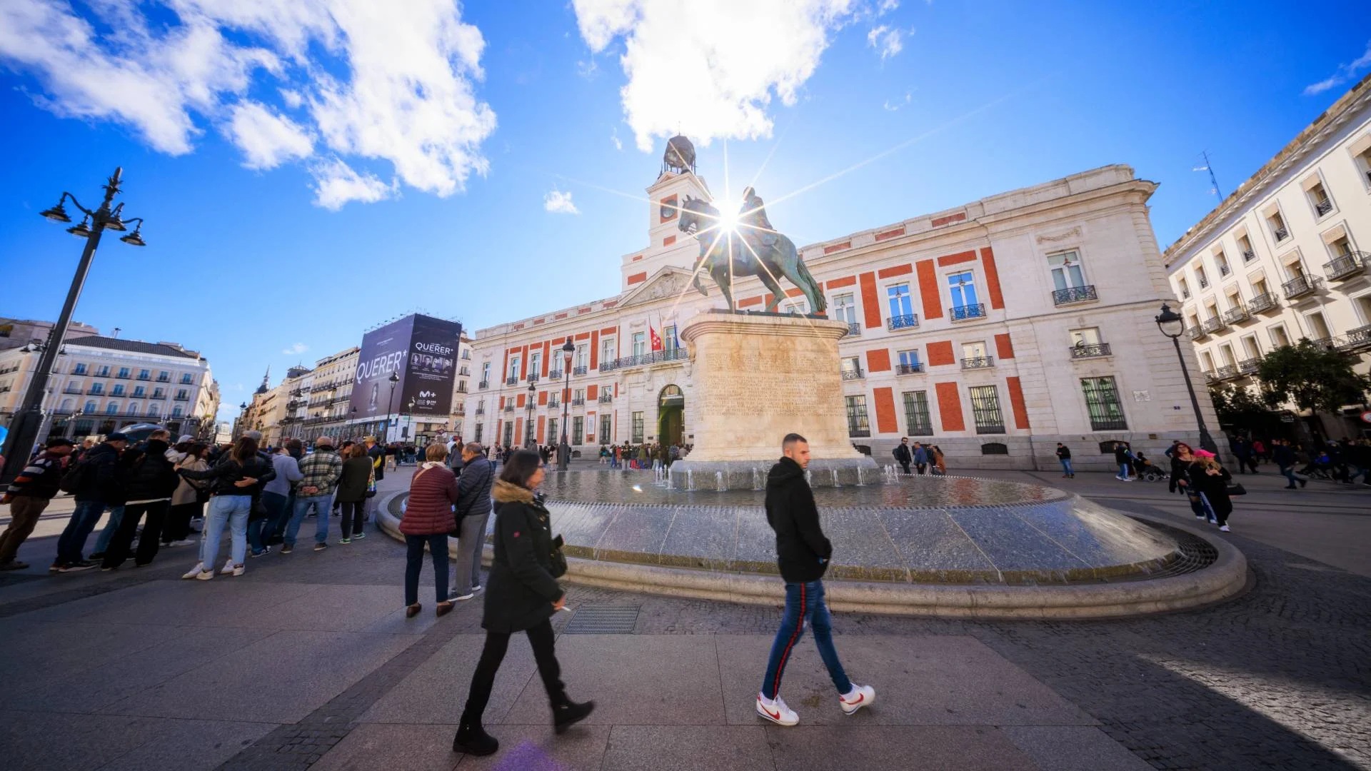 People gather around a fountain with a statue of a man riding a horse in a sunny plaza. There is a large red-and-white building with a clock tower in the background and a bright blue sky above.