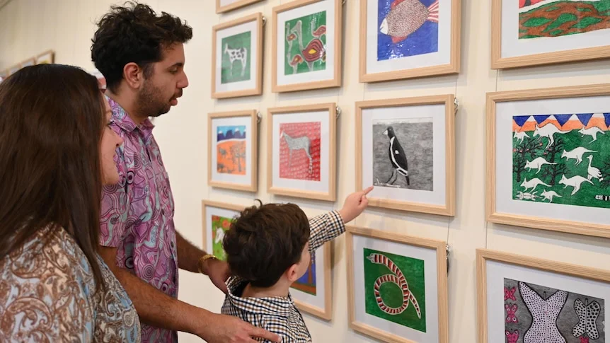 A young person points to some art on a wall, watched by a family.