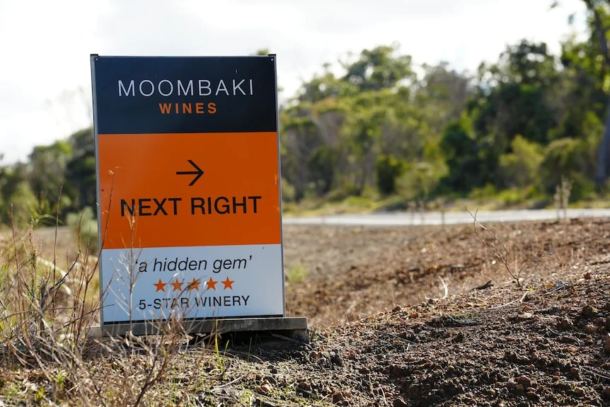a sign in on the side of a road pointing to a business, "Moombaki wines, next right".