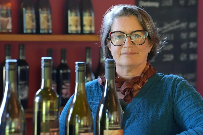 A woman stands behind a bar with wine bottles in the shot.