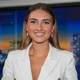A journalist standing inside a television news studio, she is wearing a white blazer and smiling at the camera.