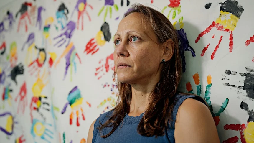 A woman, looking serious, leaning against a white wall covered in children's painted handprints in bright colours.