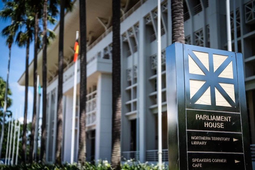 A sign that reads Parliament House on the right, with the NT parliament house behind and an Aboriginal flag displayed.