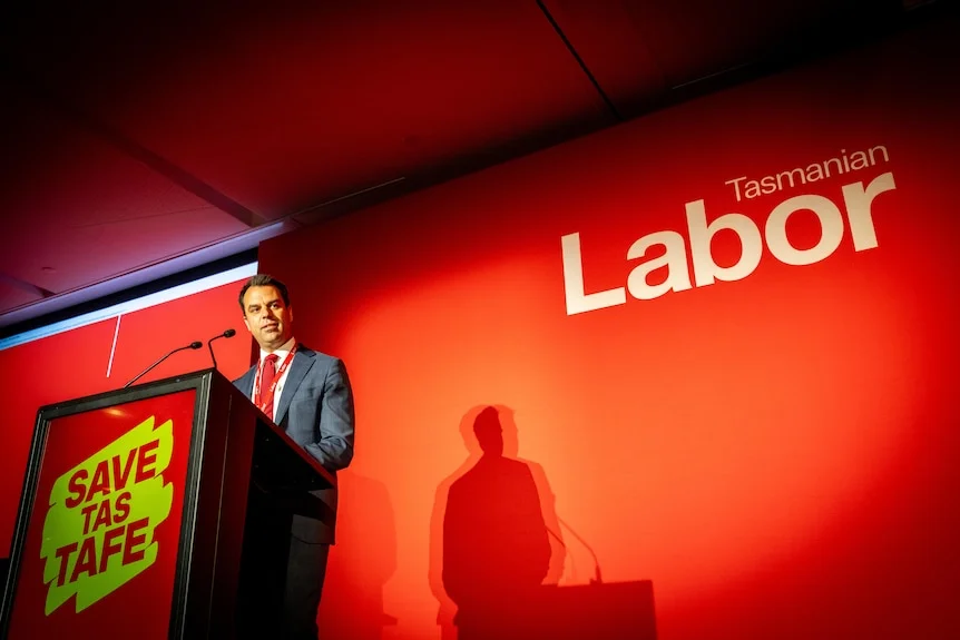 A man in a suit stands in front of a red background