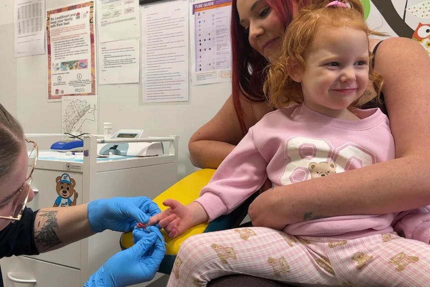 A toddler in pink on her mum's lap smiling as a nurse tests the blood on her finger