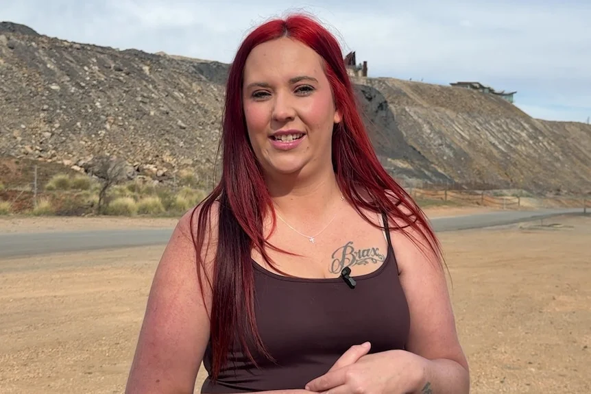 A woman with dyed red hair stands in front of a large mining lode