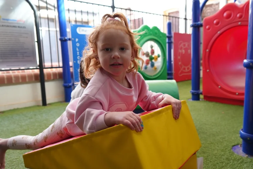 Little girl wearing pink holds onto yellow play block in playground.