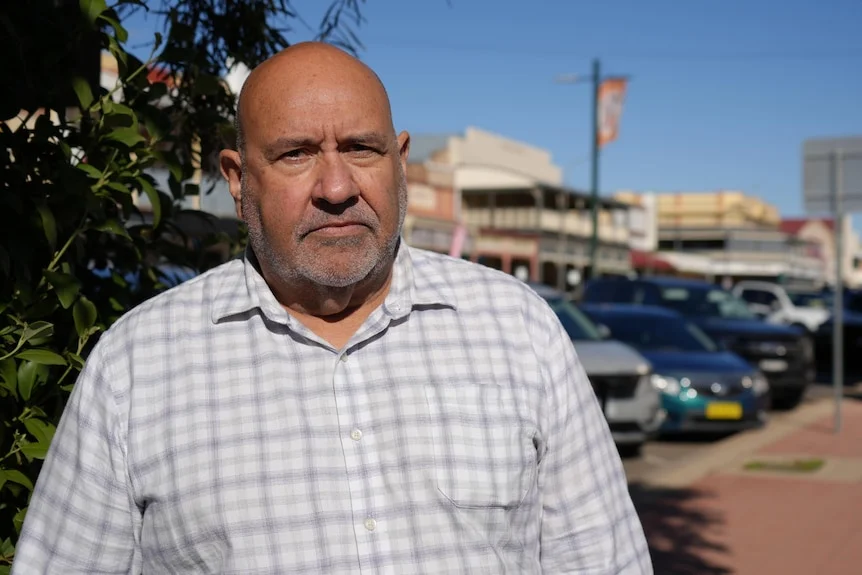 A man wearing a collared shirt, looking at the camera, standing on a sidewalk in front of cars and buildings in the background