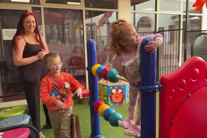 A woman in a small playground with two young children on play equipment