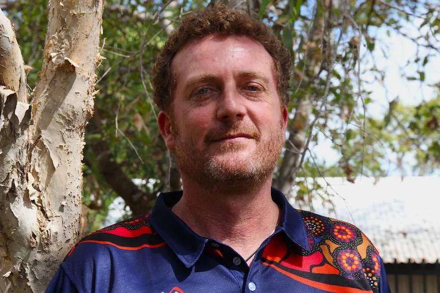 A man stands next to a tree. He wears a blue shirt with a red pattern.
