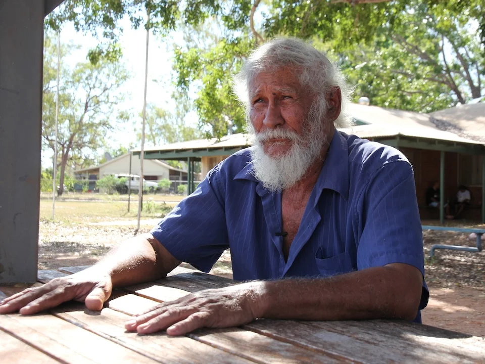 A man with white hair with his palm on the table looking to the side.