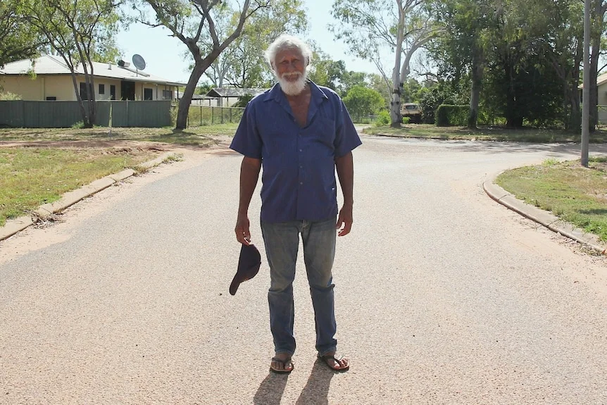 A man in a blue shirt and jeans, holding a cap in one hand, stands on a road with houses in the background.