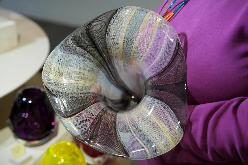 A woman with short grey hair leans over to cup her hands around a glass artwork modelled after an Indigenous eel trap.