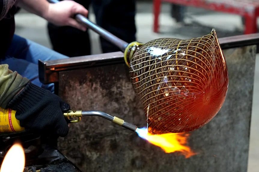A person's hands working on a woven-patterned glass sculpture with a blowtorch.