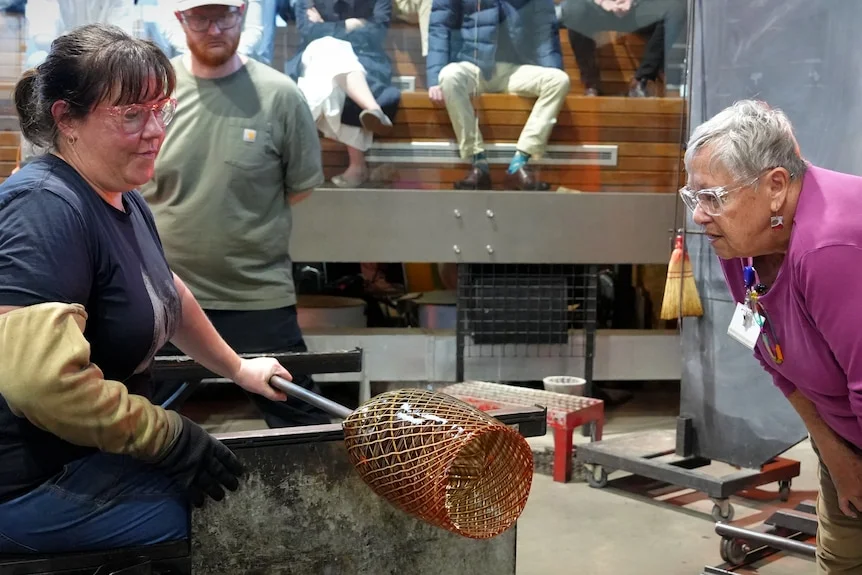 A woman with dark hair in a ponytail working on a glass sculpture as another woman supervises.