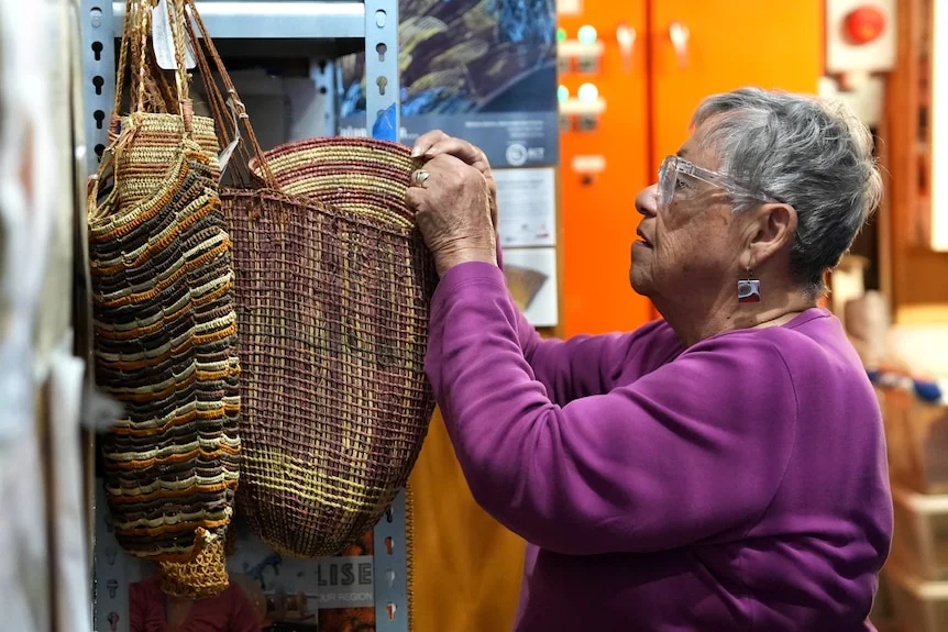 A woman with short grey hair stands in a studio looking at the weaving of an handmade bag.