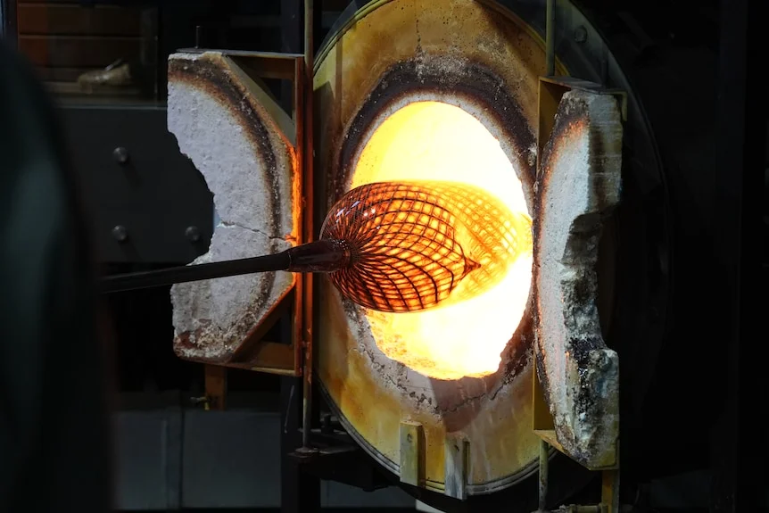 A woven-pattern glass vase attached to a blowpipe being put into a burning wall furnace.