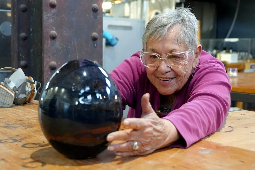 A woman with short grey hair leans over to cup her hands around a glass artwork of a large rainbow egg.