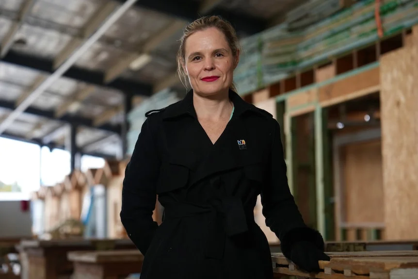 A woman with blonde hair in a ponytail stands smiling lightly outside a partially constructed home.
