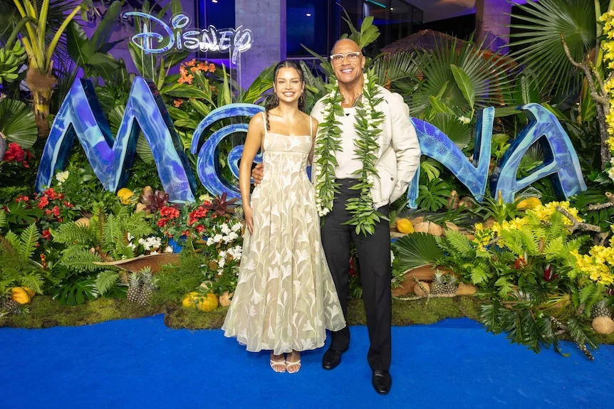 Catherine Lagaʻaia and Dwayne Johnson standing in front of a Moana sign with flowers and plants surrounding them