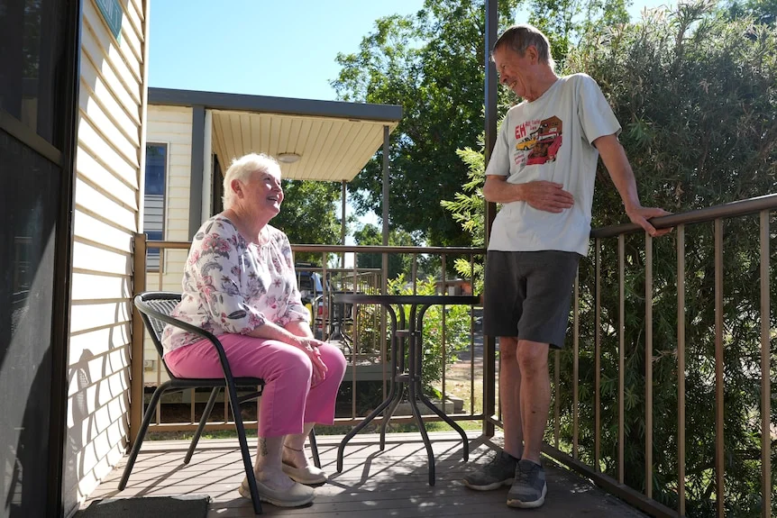 Two grey nomads on a bungalow deck, woman sitting, man standing, looking at each other, smiling.