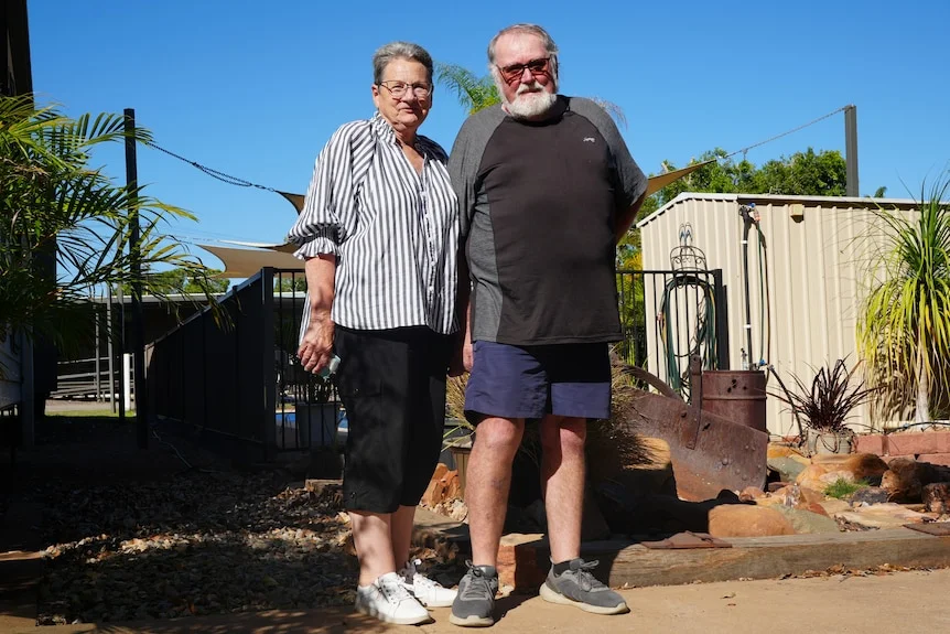 A grey nomad couple stand in front of a fence at a caravan park. They have their arms around each other and smile.