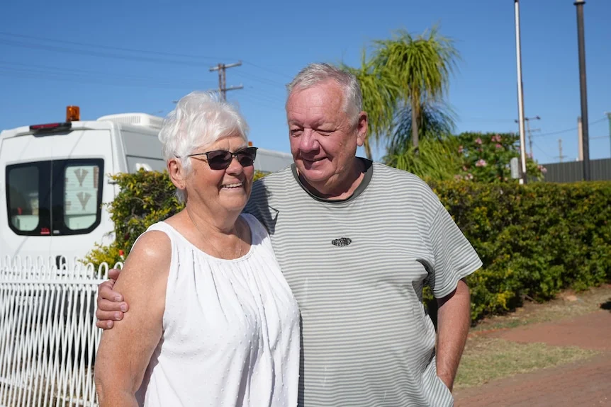 Two grey nomads stand in front of a caravan in Mount Isa and smile. They have their arms around each other.