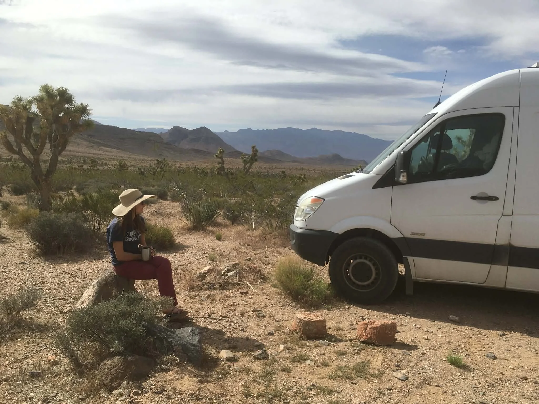 A person in a wide-brimmed hat sits on a rock in a desert landscape, embracing van life as they face a white van parked on the dirt. Mountains and Joshua trees stretch across the background.