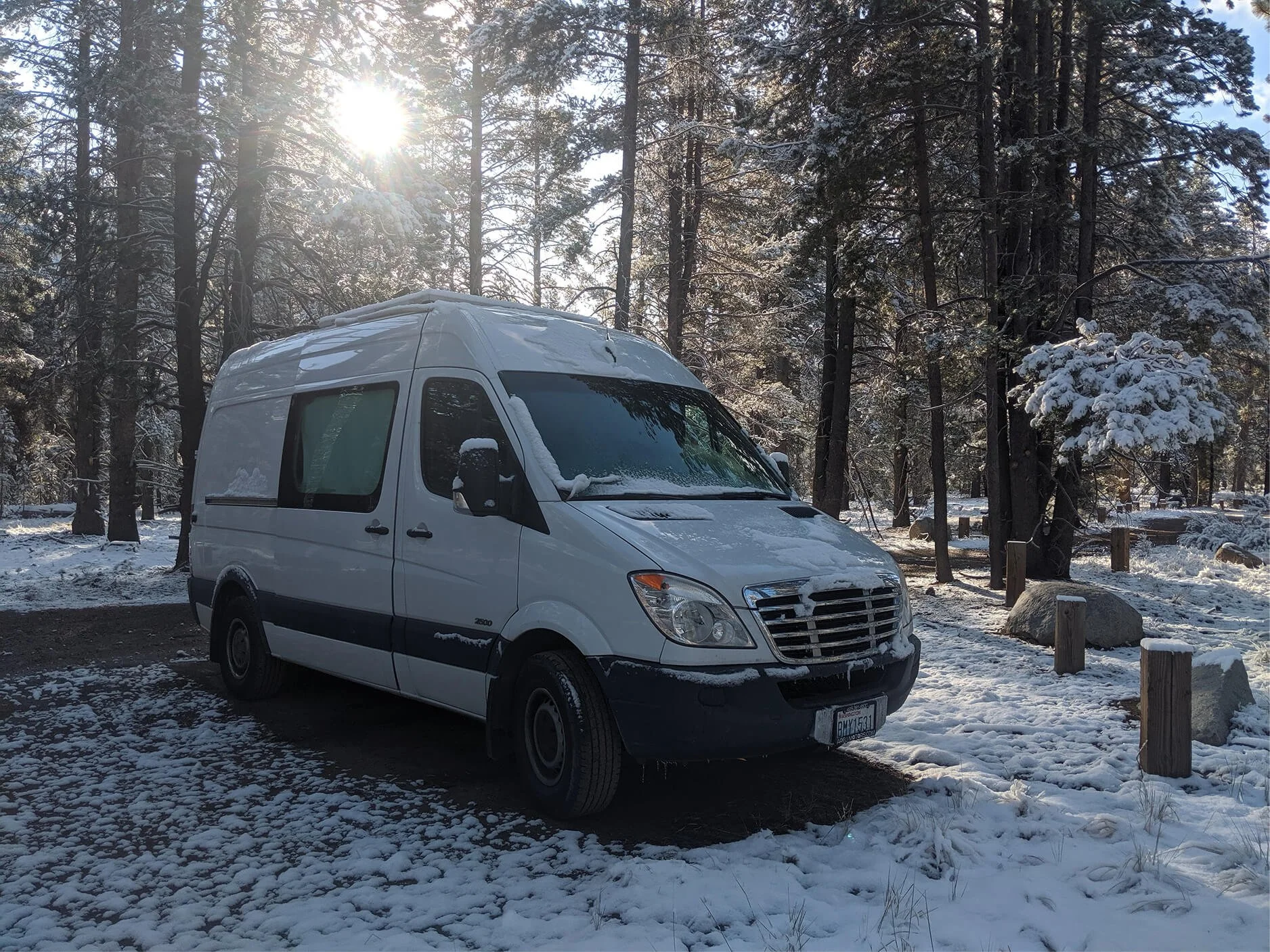 A white van, perfect for van life, is parked in a snowy forest clearing with sunlight filtering through tall trees in the background.
