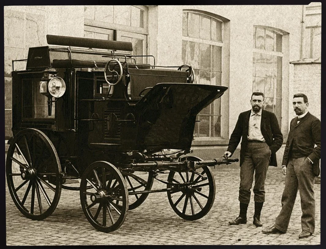 Two men stand beside a vintage D'Ieteren motor vehicle with large wooden wheels, parked on a cobblestone surface in front of a building with large windows.