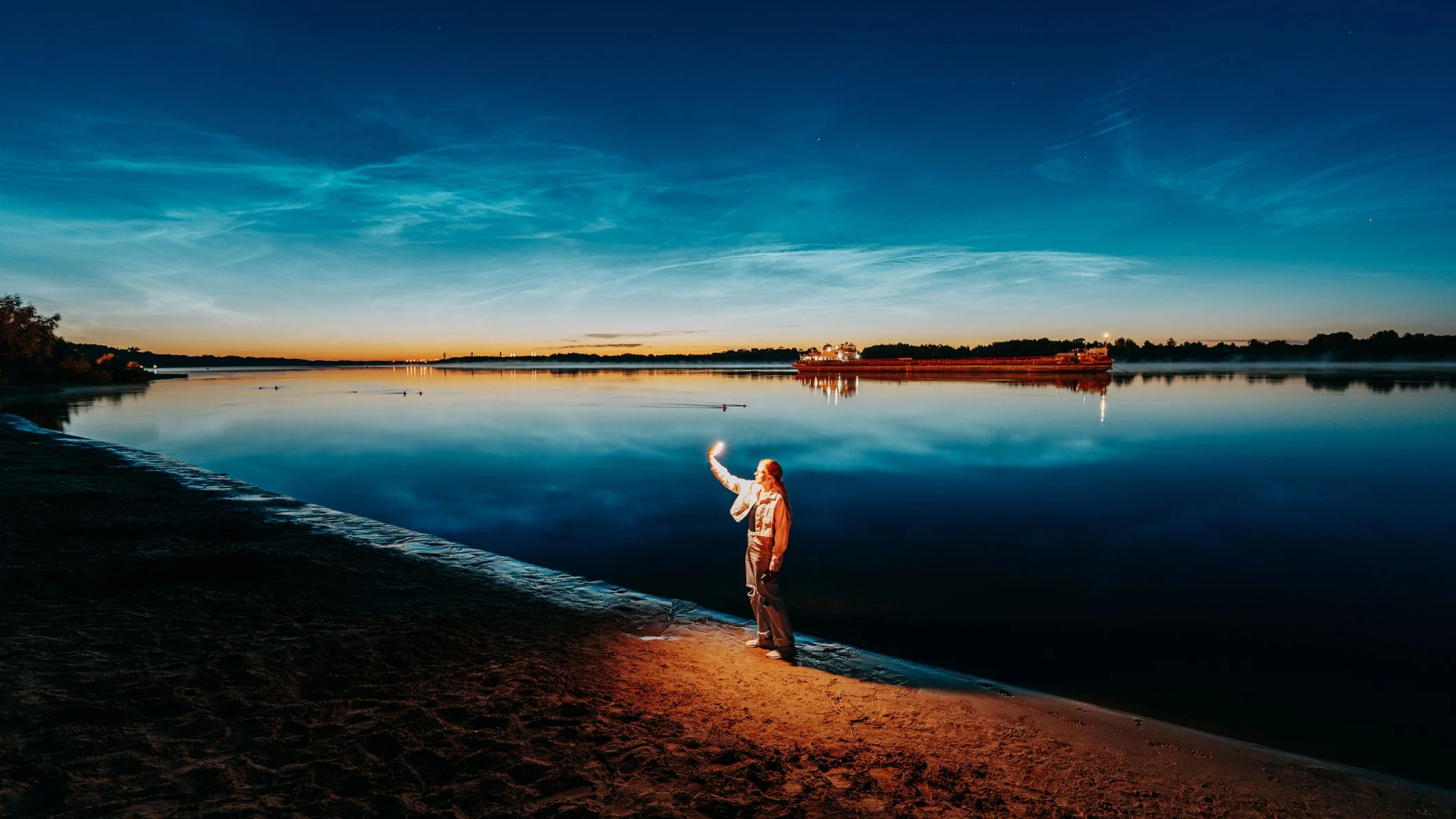 Woman takes pictures with smartphone of noctilucent clouds on the river bank at night