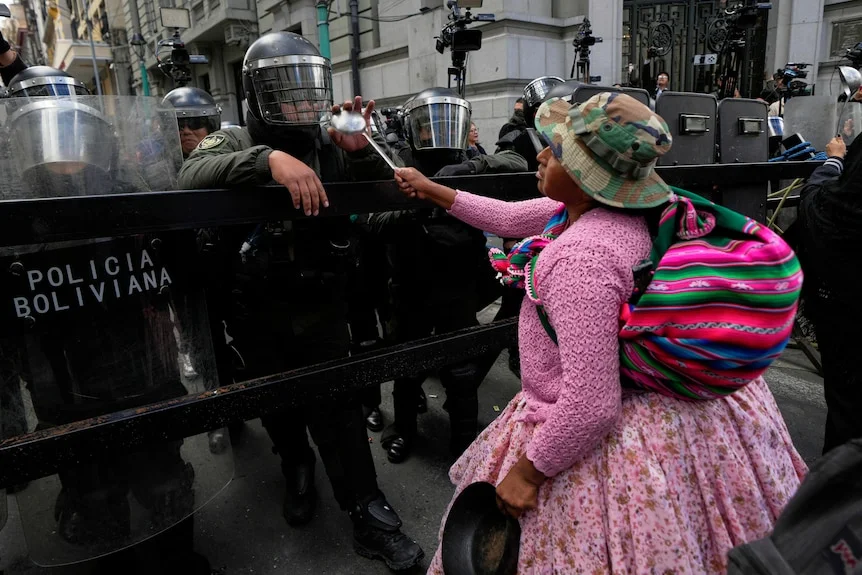 A woman in pink with a colourful traditional bag slung over shoulder holds a ladle at a group of police in riot gear.
